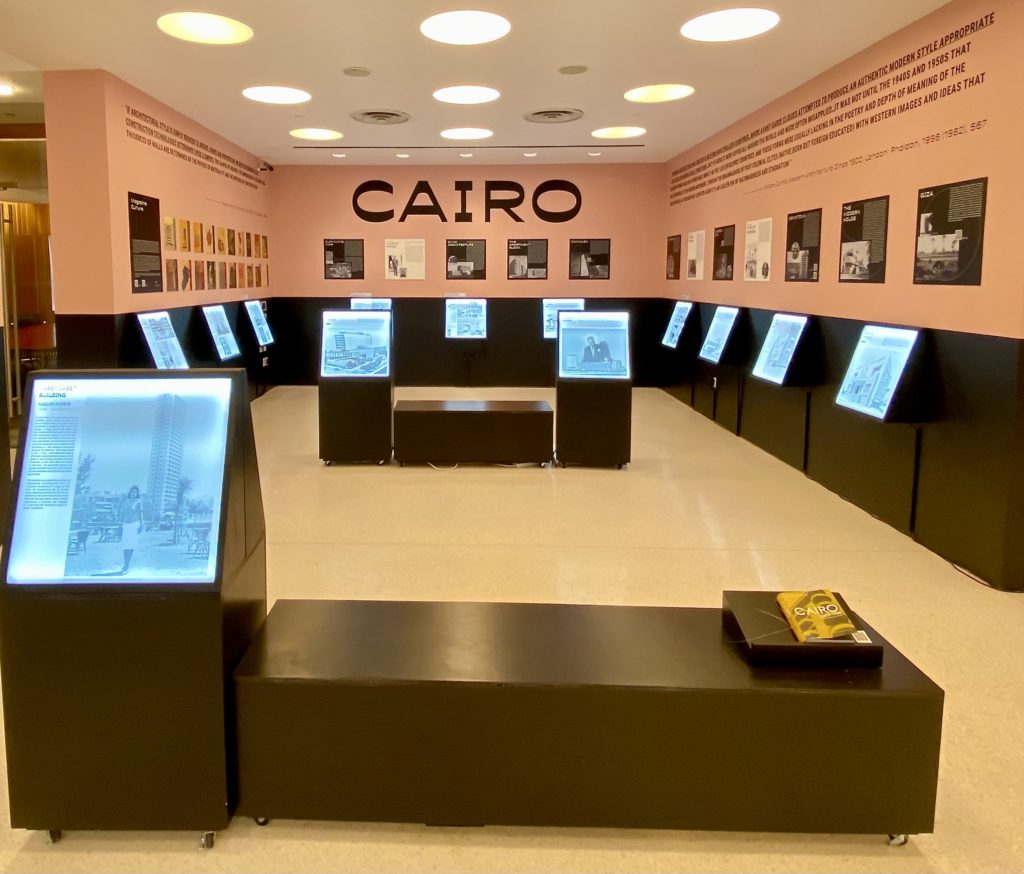 The first-floor exhibition space, photographed from its entrance, has pink walls and black-cased lightbox panels with white text and black-and-white backlit images of Modernist works of Egyptian architecture and urban planning.